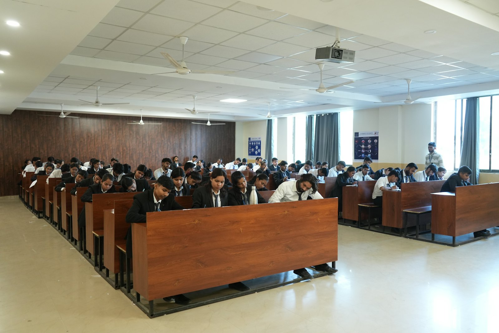 Students writing an exam in a large hall while an invigilator stands at the side, outlined by the AI camera.